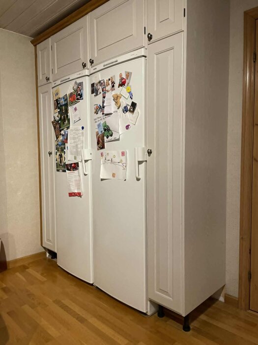 Vita kylskåp och frysskåp med skåp ovanpå och på sidorna, fastsatta i väggen utan synliga skenor, stående på trefotade ben på parkettgolv.