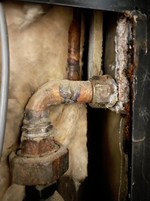 Rostig kopparröranslutning på värmeväxlare som läcker, omgiven av isolering och metalldelar.