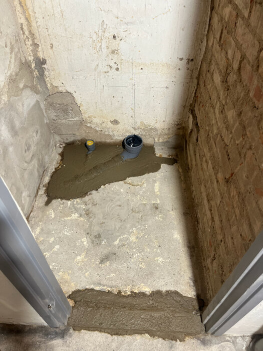 Cementerat hörn med rörläggning, omgiven av ojämna murväggar. Förberedelse för renovering med planerad klinker och tapet.