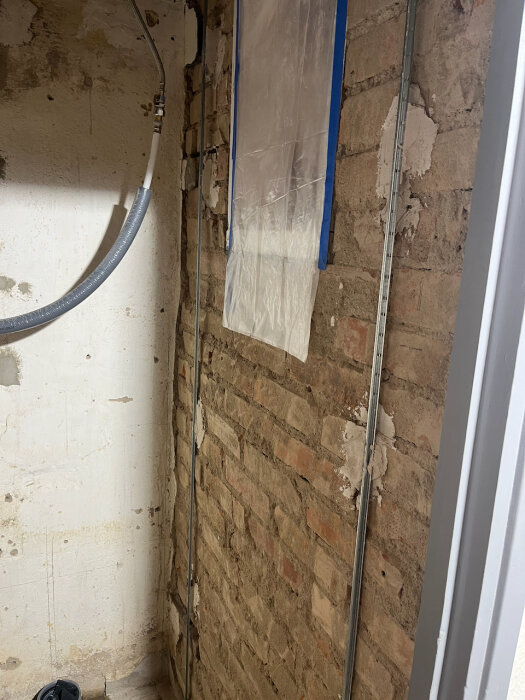 Renovering av gammal tegelvägg med delvis putsning, rördragning och plastskydd på väggen, förberett för klinker och tapet i badrum.