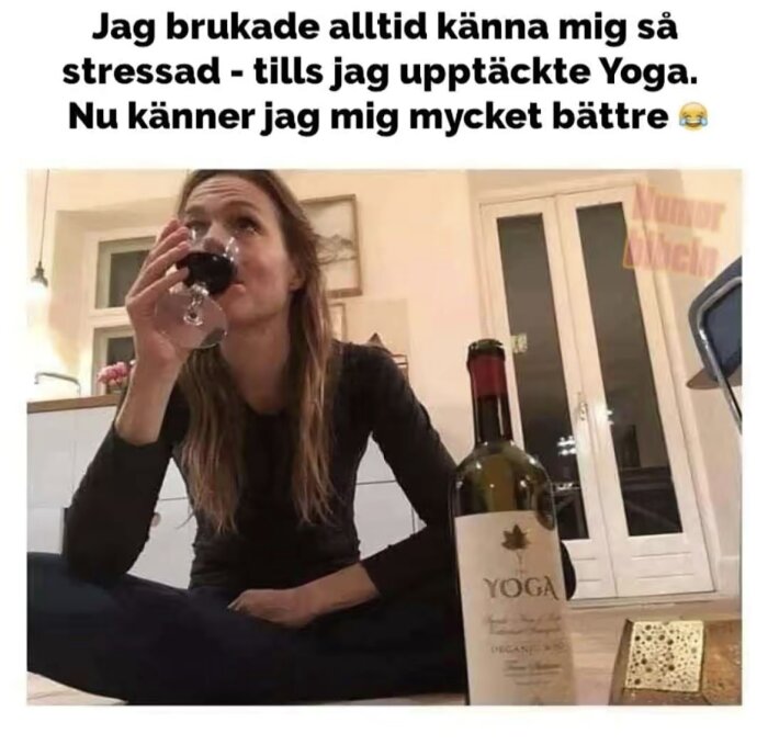 Kvinna sitter på golvet, dricker rödvin från ett glas. En vinflaska med etiketten "Yoga" står framför henne.