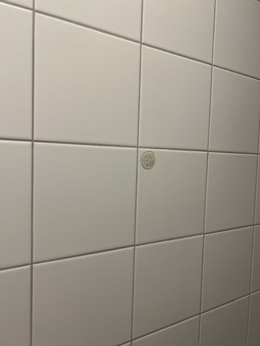 Vitt kakel i badrum med en rund öppning där plastprodukt tagits bort, lämnar limrester. Möjligt förberett för elinstallation, t.ex. handdukstork.
