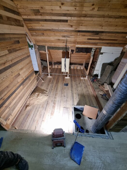 Under renovering av ett badrum, med blottat bjälklag och isolering synlig, samt en hålighet i golvet och verktyg på betongytan.