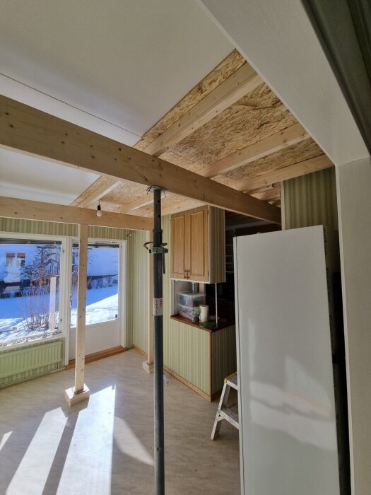 Rum under renovering med nytt träbjälklag och stödregel mot taket, sol skiner genom fönstret, stege och verktyg i hörnet.