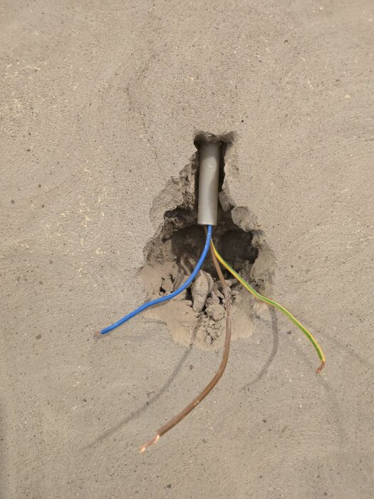 Elektrisk kabelinstallation i en vägg med synliga, oskyddade kablar i olika färger som sticker ut från ett hål i betong.