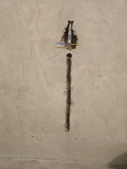 En vägg med en vertikal ränna och synliga elledningar samt hål för ett eluttag, betongvägg med sprickor.
