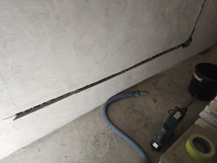 Vägg med utfräst spår för eldragning, dammsugarslang på golvet och arbetshjälpmedel bredvid, inklusive borrmaskin och tejp.