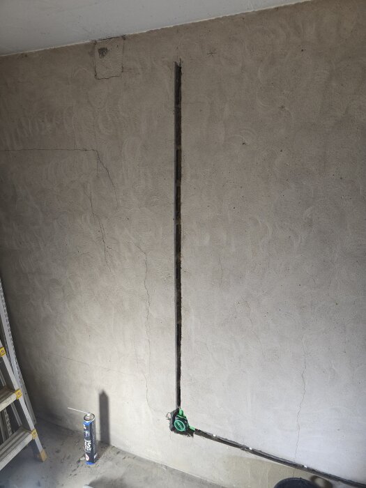 Vägg med utskuren kanal för elrör, gröna infällda dosor och en sprayburk på golvet. Sprickor syns på murens yta. Stege står intill.