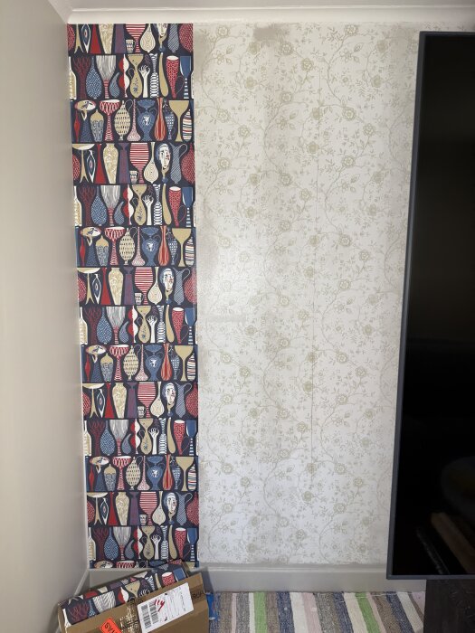 Två olika tapeter, en med färgglada vaser och en med blommönster, möts i ett hörn av ett rum. Kartonger står på golvet.