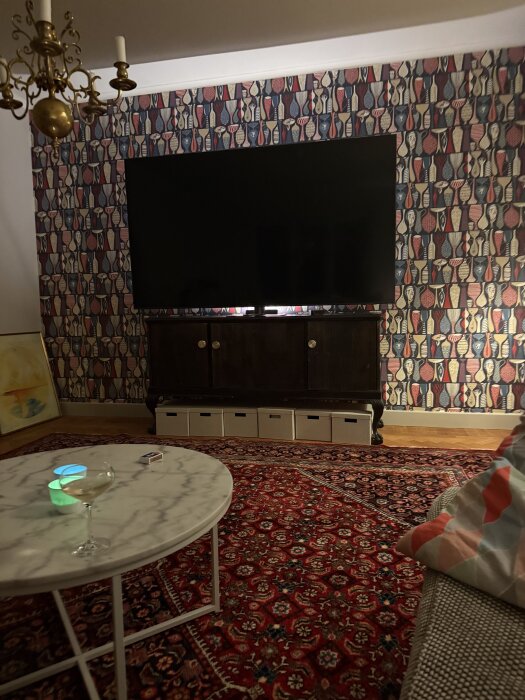 Vardagsrum med stor TV på mönstrad tapetvägg, rött orientaliskt matta, marmorbord med dryck och soffkudde med färgglatt mönster.