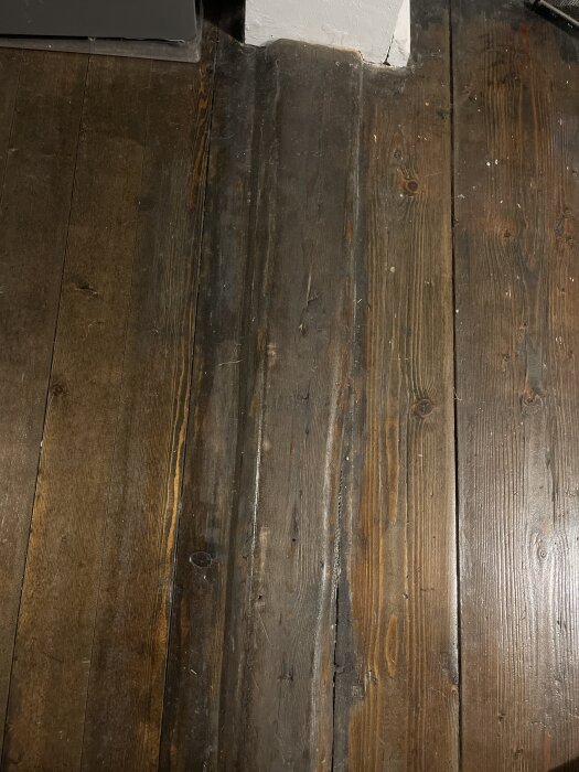 Tröskel mellan två rum där golvplankor i mörkt trä syns. Dagljus kommer in från ett fönster och skapar ljusreflektioner på golvet.