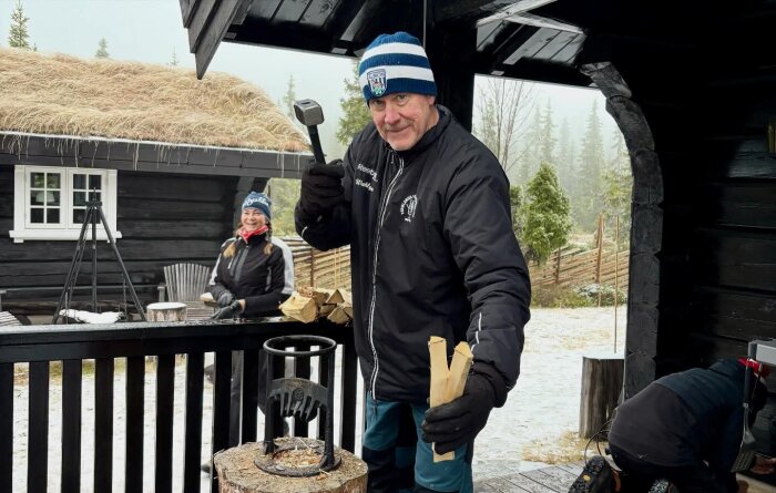 Man klädd i vinterkläder håller en yxa och tändved utomhus framför en stuga med torvtak, en kvinna ler i bakgrunden. Någon arbetar nära marken.