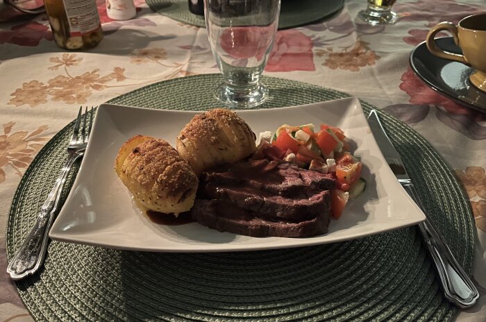 Flankstek med vitlöksmarinad, hasselbackspotatis med timjansmör, rödvinssås och grönsaker på tallrik, dukat med bestick.