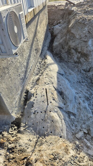 Hålad bergknalle nära husgrund för förberedelse av snigeldynamitbrytning, med borrhål i rader och ett ventilationsaggregat på väggen.