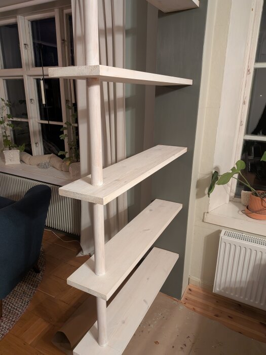 Öppen rumsavdelare med vita hyllplan och runda stolpar mot en grön vägg, synliga springor mellan hyllorna och väggen, inomhusmiljö.