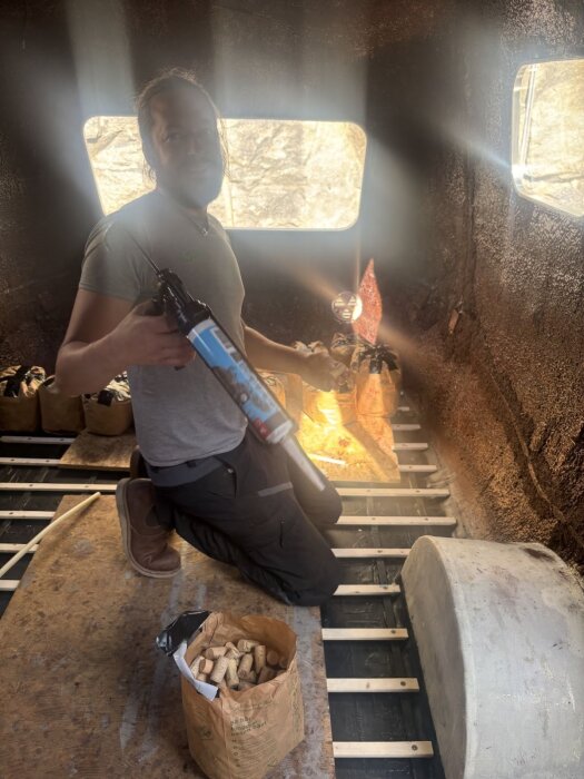 Person limmar vinkorkar på vägg i en husvagn, håller en silikonpistol. Ljuset strömmar in från fönster, påsar med korkar syns i bakgrunden.