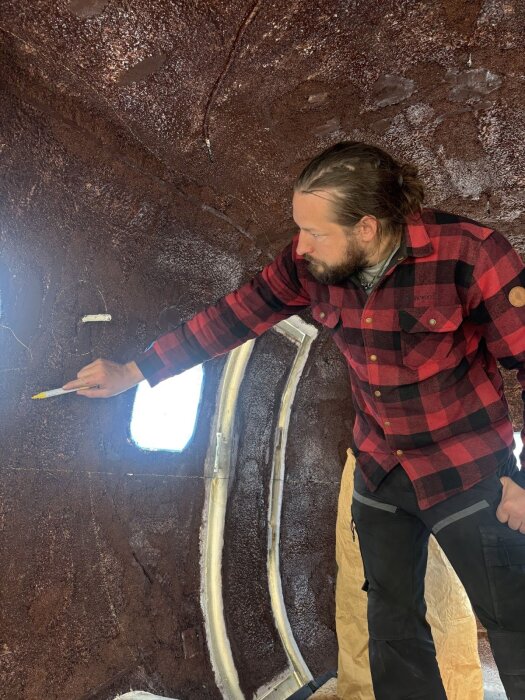 Man i rutig skjorta skissar på ett mönster på en kupad, brun vägg med en markerpenna, förberedande för korkprojektet.