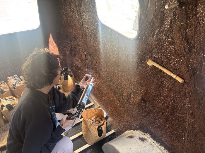 Person limmar vinkorkar på en mörk vägg med hjälp av en tublim. Ljuset skiner in genom ett fönster, flertalet korkar och verktyg ses runt omkring.