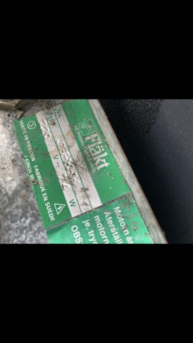 Etikett på fläktmotor med texten "Fläkt", tillverkad i Sverige, samt teknisk information. Smutsig yta visar slitage.