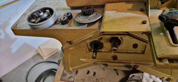 Maskinens interiör visar utspridda kugghjul och delar, inklusive en delvis isärplockad rund växellåda och olika skruvar på arbetsbänken.