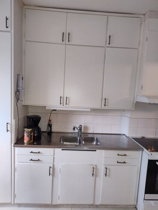 Vitt kök med över- och underskåp, diskho och kakelplattor. Synliga köksapparater och vägguttag på sidan. Del av renoveringsdiskussion.