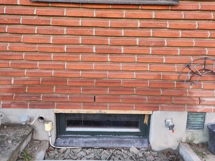 Röd tegelfasad med ett källarfönster delvis omgivet av trä. En kran till höger och stentrappa till vänster.