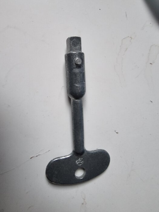 Nyckel till vindslucka med metallhandtag och fyrkantsfattning liggande på vit bakgrund.