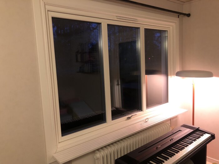Fönster med mörk utsikt, bredvid en golvlampa och ett elpiano på en vitvägg.