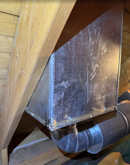 Metallåda i ett gammalt trähus, inneslutande en frånluftsfläkt kopplad till ett ventilationsrör; bilden visar hörnet av lådan och röret.