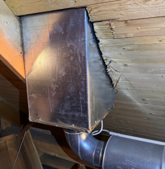 Ventilationslåda med plåtskal vid tak i äldre hus; skadad träpanel intill, del av ventilationsrör synligt nedanför.