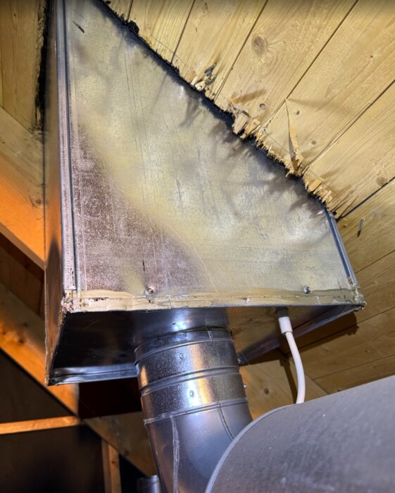 Ventilationsbox med rund kanalanslutning och träram i vad som verkar vara ett takutrymme. Boxen ser sluten ut och har en synlig kabel bredvid.