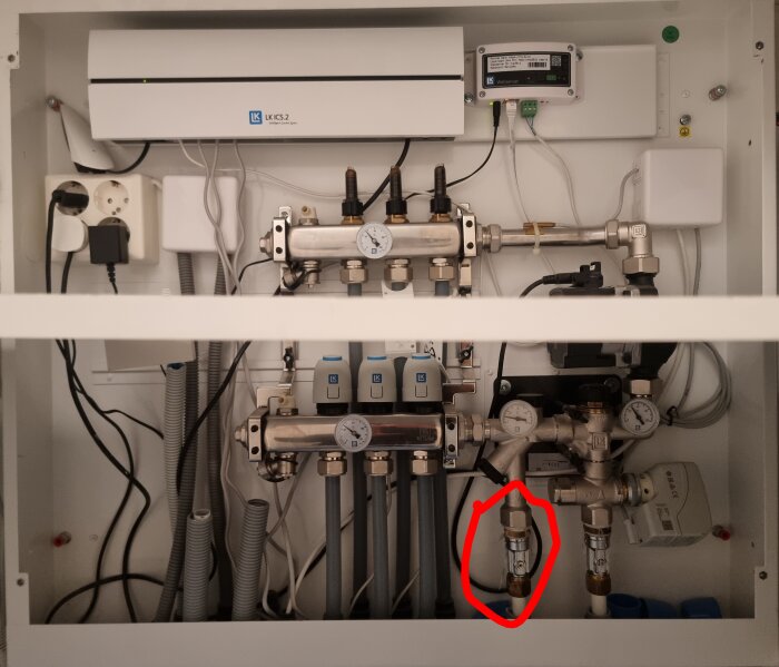 Bild av ett golvvärmesystem med rör och termometrar, fokus på ett inringat rör som misstänks påverka värmen. Hylla syns på bilden.