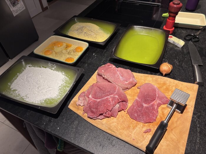 Rå skivad fläskkött på en skärbräda med en köttbankare. Omkring ligger mjöl, ägg och brödsmulor.