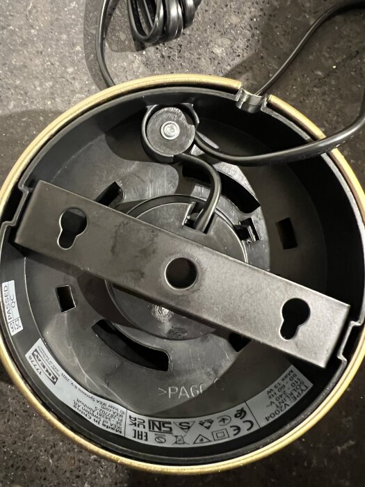 Rund taklampa från IKEA med svart kabel, metallfäste och synliga etiketter med specifikationer och säkerhetssymboler.