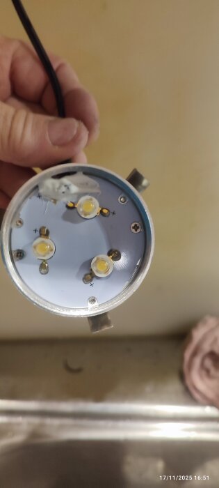 Hand håller i en trasig LED-skåpslampa med synliga kretsar och ledningar, ovanför en diskbänk.