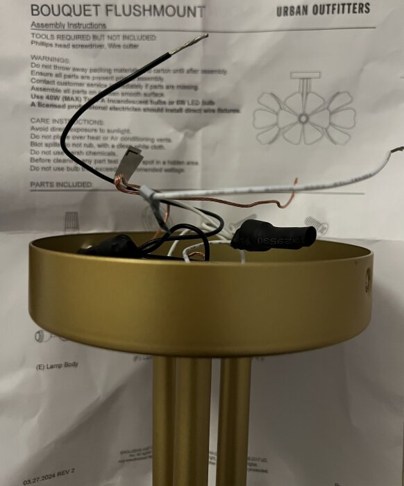 Guldfärgad taklampa med synliga kablar, placerad framför monteringsinstruktioner och varningar av märket Urban Outfitters.