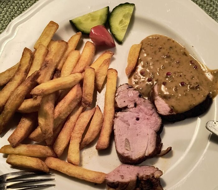 Fläskfilé med rosépepparsås, pommes frites, gurka och röd paprika på en vit tallrik.