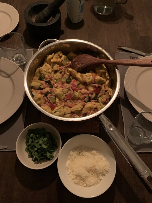 En kastrull med bacontortellini, omgiven av skålar med riven parmesan och färsk basilika på ett dukat bord.