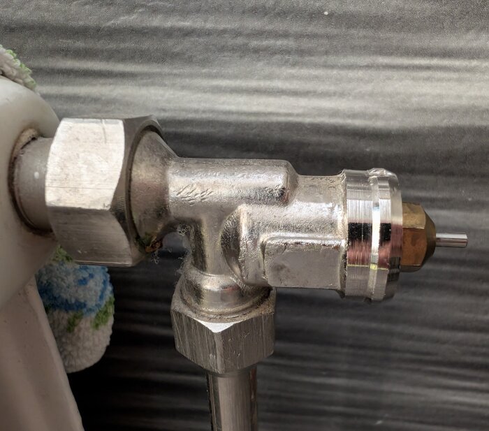 Närbild av en metallisk radiatorventil från sidan, ansluten till ett rör. Ventilen är av obestämd typ och visas mot en mörk bakgrund.