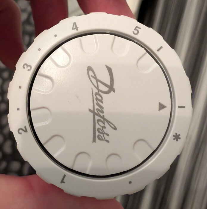 Vit Danfoss-termostat med temperaturinställningar från 1 till 5, hålls i en hand, mot en suddig bakgrund.