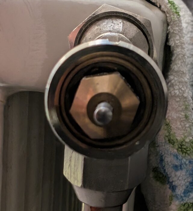 Närbild av en radiatorventil i metall med hexformad mittdel, omgiven av en smal ring och delvis täckt av en vit handduk.