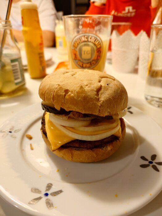 Hamburgare med gul senap, ost, dubbel lök, saltgurka, ketchup och sourcreme på en tallrik. Bakgrunden visar drycker och ketchupflaska.