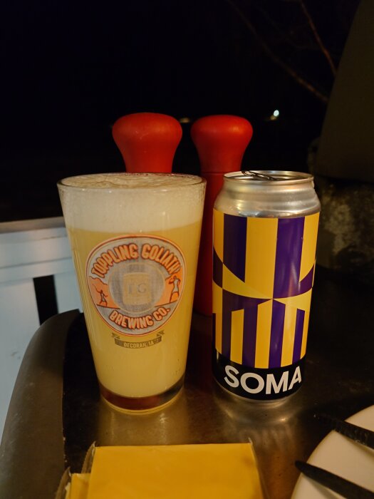 Ett ölglas med Toppling Goliath-logga och en burk med texten "SOMA" står bredvid varandra på ett bord, med röd pepparkvarn i bakgrunden.