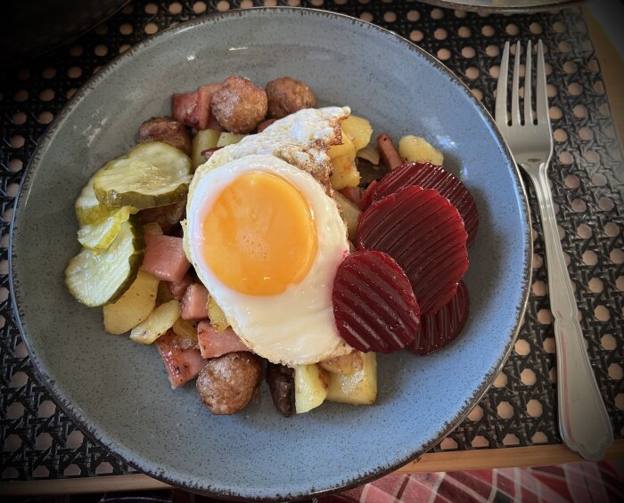 Tallrik med pyttipanna, köttbullar, falukorv, potatis, inlagda rödbetor, gurka och stekt ägg, serverat på ett bord.