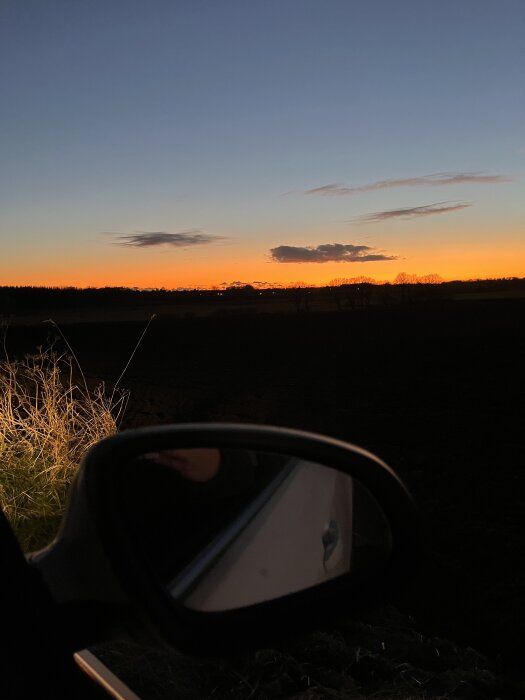 Utsikt från bilens sidofönster av en solnedgång över ett fält med en klar himmel och några moln.