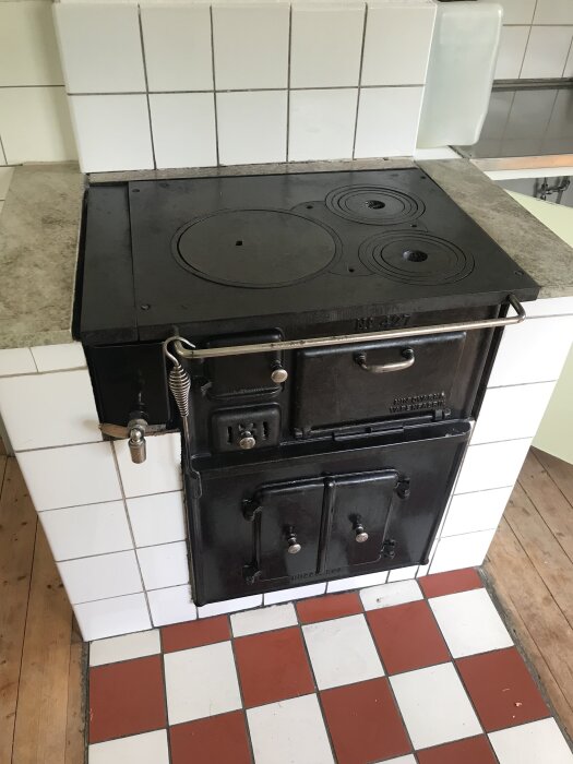 Gjutjärnsvedspis med smala eldningsluckor, stående i ett kök med vit kakel och rödvit rutigt golv.