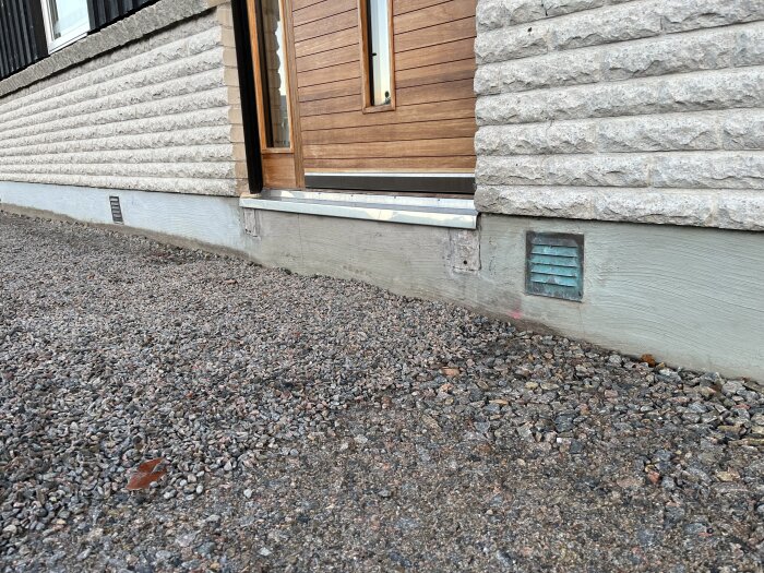 Husets fasad med entrédörr, stengrund och ventil. Framför ligger grus. En elslang syns klämd under en metallplåt vid trottoarkanten.