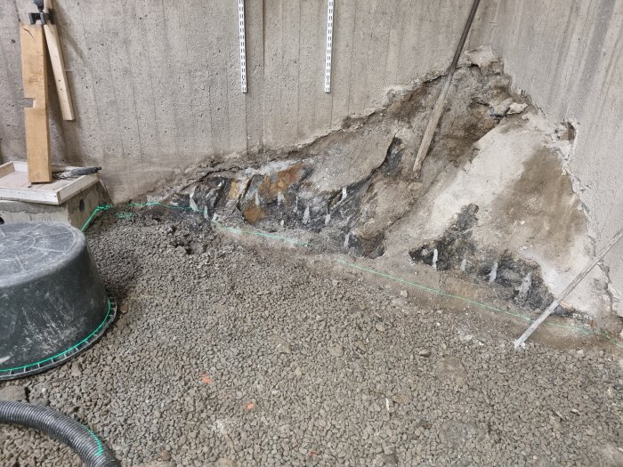 Byggrenovering med en betongvägg och exponerad grund, omgiven av grus och byggmaterial som trä och rör.