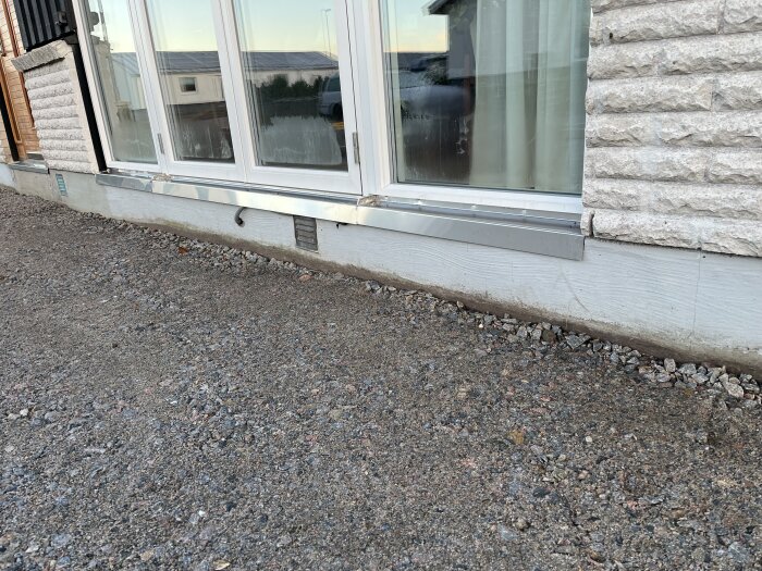 Fasad med glasparti och grus framför, synlig rostfri plåt och elkabel klämd under plåten. Förberett för planerat altanbygge.