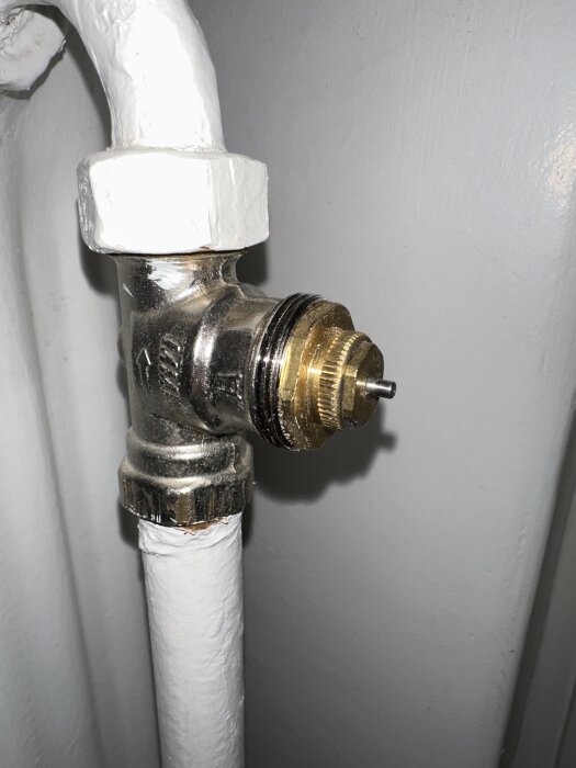 Gammal termostatanslutning med metallgängor på ett vitt rör, del av ett värmesystem.